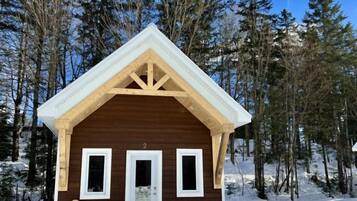 A small wooden cabin with a gabled roof, a door, and two windows, surrounded by snow and trees.