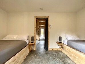 Een kamer met twee bedden, een houten vloer, een deur naar een gang en een lichtschakelaar aan de muur.