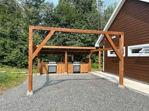 An outdoor barbecue area with a wooden pergola structure, two barbecue grills, and a building with a brown siding exterior. - Mini-Chalet #3 – Near the River (Saint-Gabriel-de-Valcartier)