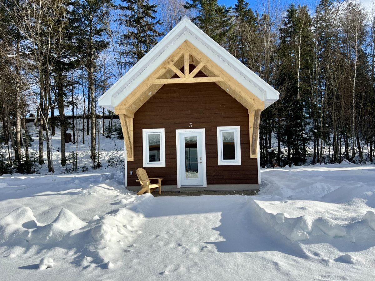 Une petite cabane en bois, avec une porte et des fenêtres blanches, entourée de neige et d’arbres.