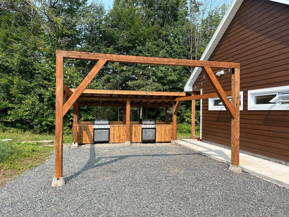 A wooden pergola structure with barbecue grills underneath, set against a building with a brown siding exterior.