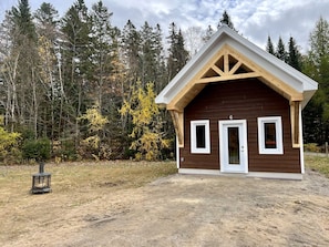 A small wooden cabin with a gabled roof, a door, and two windows, surrounded by a forest.