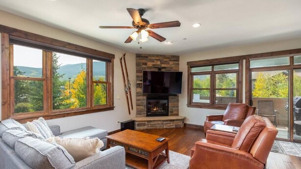 A spacious living room with a fireplace, large windows, and a ceiling fan. - Private Hot Tub, Walk to Main St, Close to Ski Resorts, Incredible Views (Firsco)