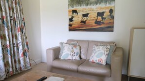 A living room with a beige sofa, floral cushions, a wooden coffee table with a remote control and a book, a painting of cows on the wall, and a curtain with a floral pattern. - Twodogfolly at Dangarsleigh, Cottage at Dangarsleigh (Armidale)