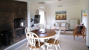 A cozy living room with a brick fireplace, a round wooden dining table with white chairs, a sofa with cushions, and a painting on the wall. - Twodogfolly at Dangarsleigh, Cottage at Dangarsleigh (Armidale)