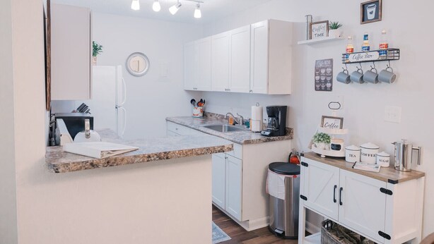 Una cocina moderna con armarios blancos, encimera de granito, espacio para café y un cubo de basura.