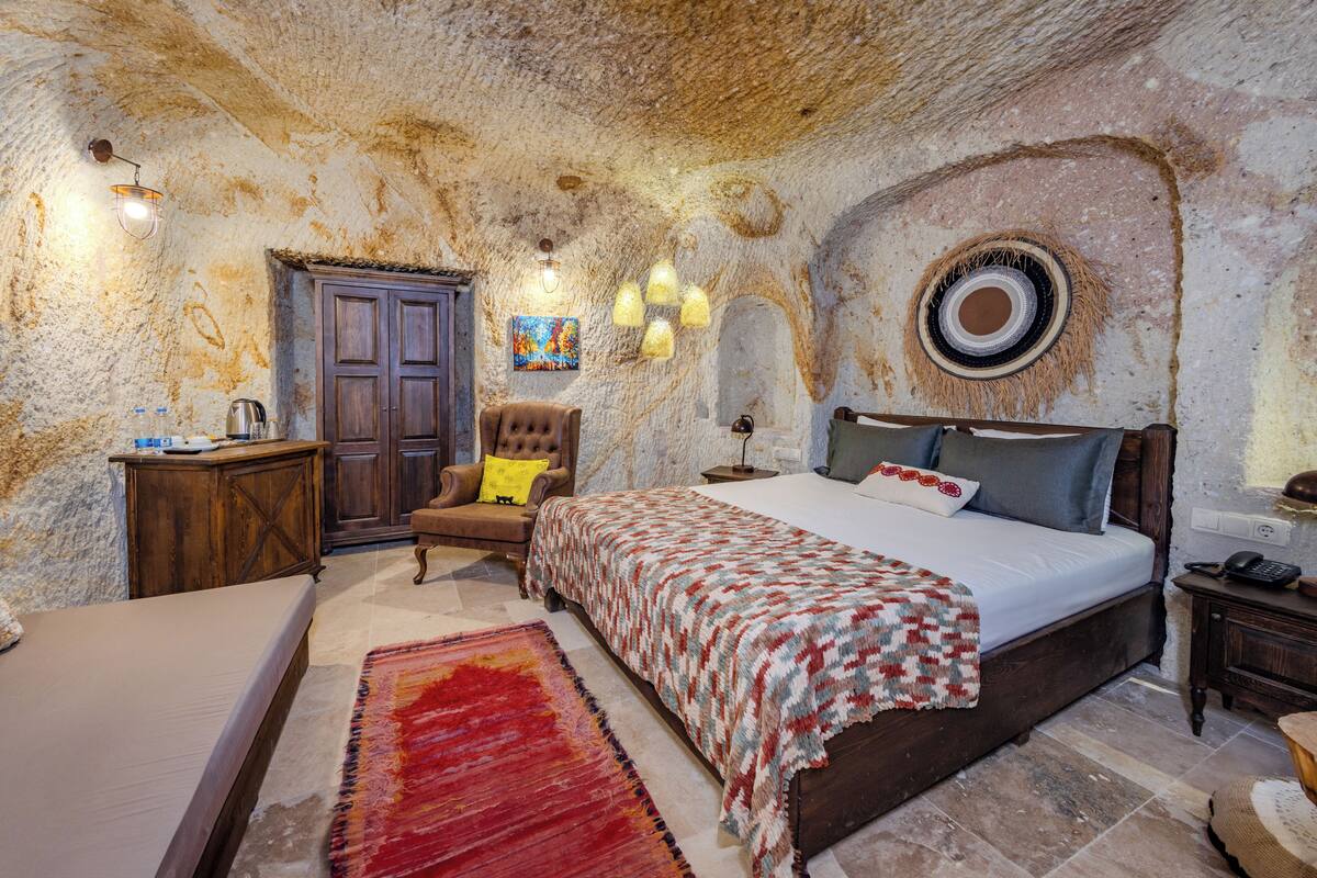 A bedroom with a rustic stone wall, a wooden bed, a red and white patterned rug, a wooden nightstand, and a chair with a yellow cushion.