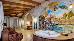 A luxurious room with a large wooden bathtub, a mural on the wall, and a cozy seating area. - Emars Cave Cappadocia (Ürgüp)