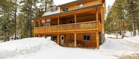 Uma cabana de tronco de dois andares com varanda, rodeada por neve e árvores.