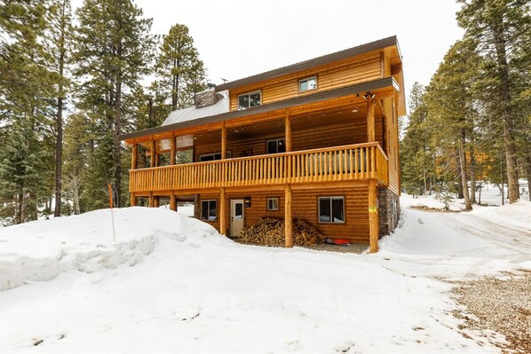 A two-story log cabin with a balcony, surrounded by snow and trees.