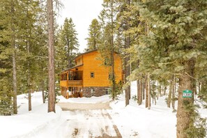 Une cabane en rondins dotée d’une terrasse en bois, entourée d’un paysage enneigé et de grands pins.
