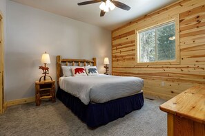 Ein Schlafzimmer mit hölzernem Kopfteil, einem Bett mit grauer Bettdecke, zwei Nachttischen mit Lampen, einem Fenster und einem Deckenventilator.