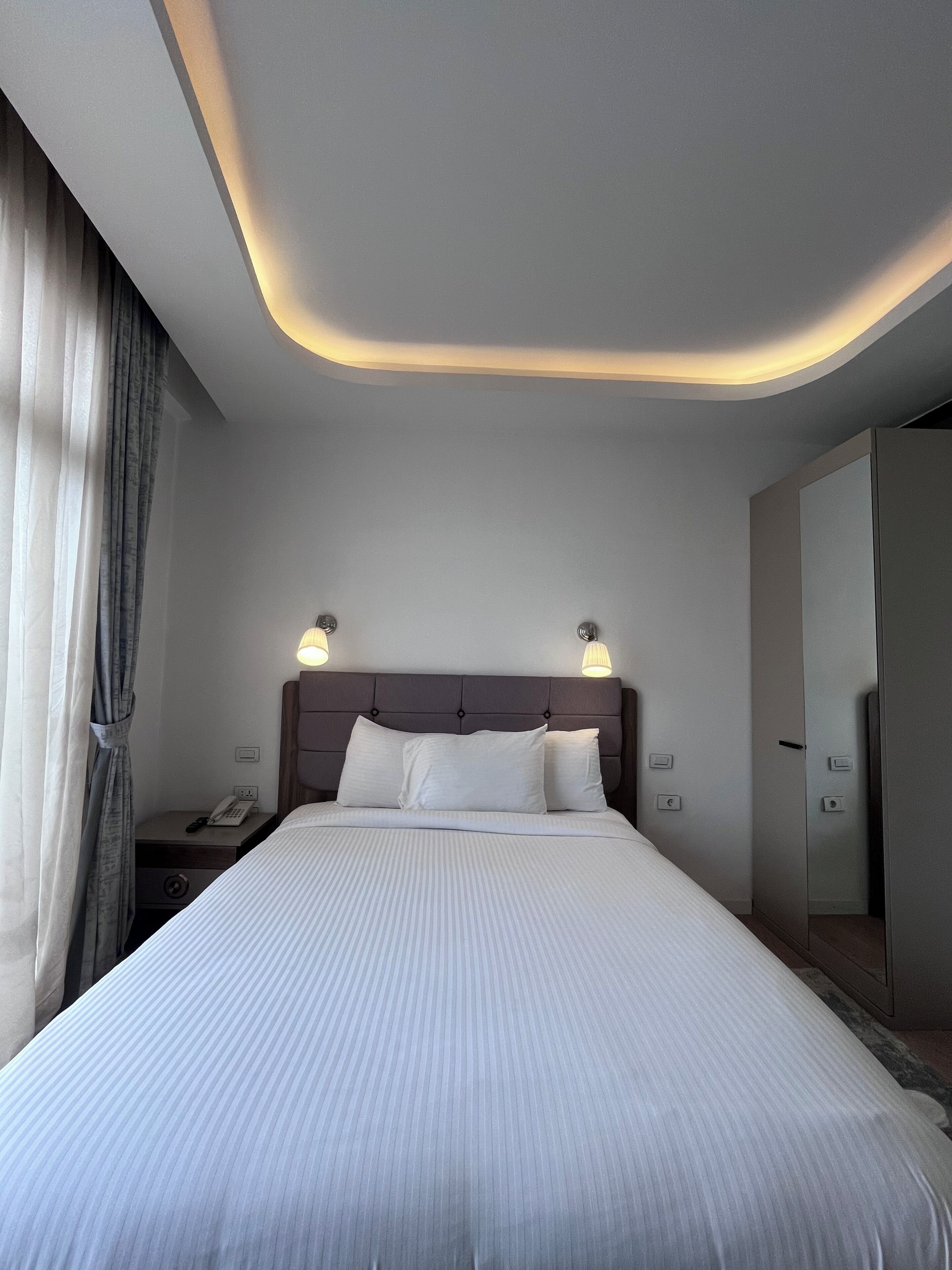 A neatly made bed with a headboard, bedside lamps, a nightstand with a phone, and a mirror on the wall.