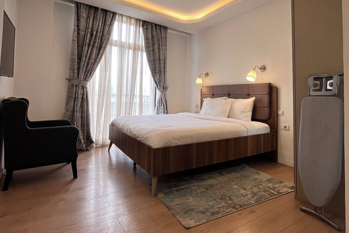 A modern bedroom with a wooden bed, a black armchair, a standing heater, and a ceiling light fixture.