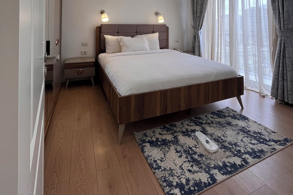 A modern bedroom with a wooden bed, bedside lamps, a large window with curtains, and a ceiling light fixture.