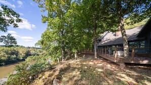 A riverside property with a wooden deck overlooking a river, surrounded by trees and a clear sky. - Exclusive Wooded Chalet w Magnificent River Views (Shepherdstown)