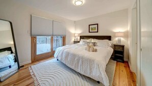 Ein Schlafzimmer mit einem großen Bett, einem Fenster mit Jalousien, einem Nachttisch mit Lampe und einem Spiegel an der Wand.