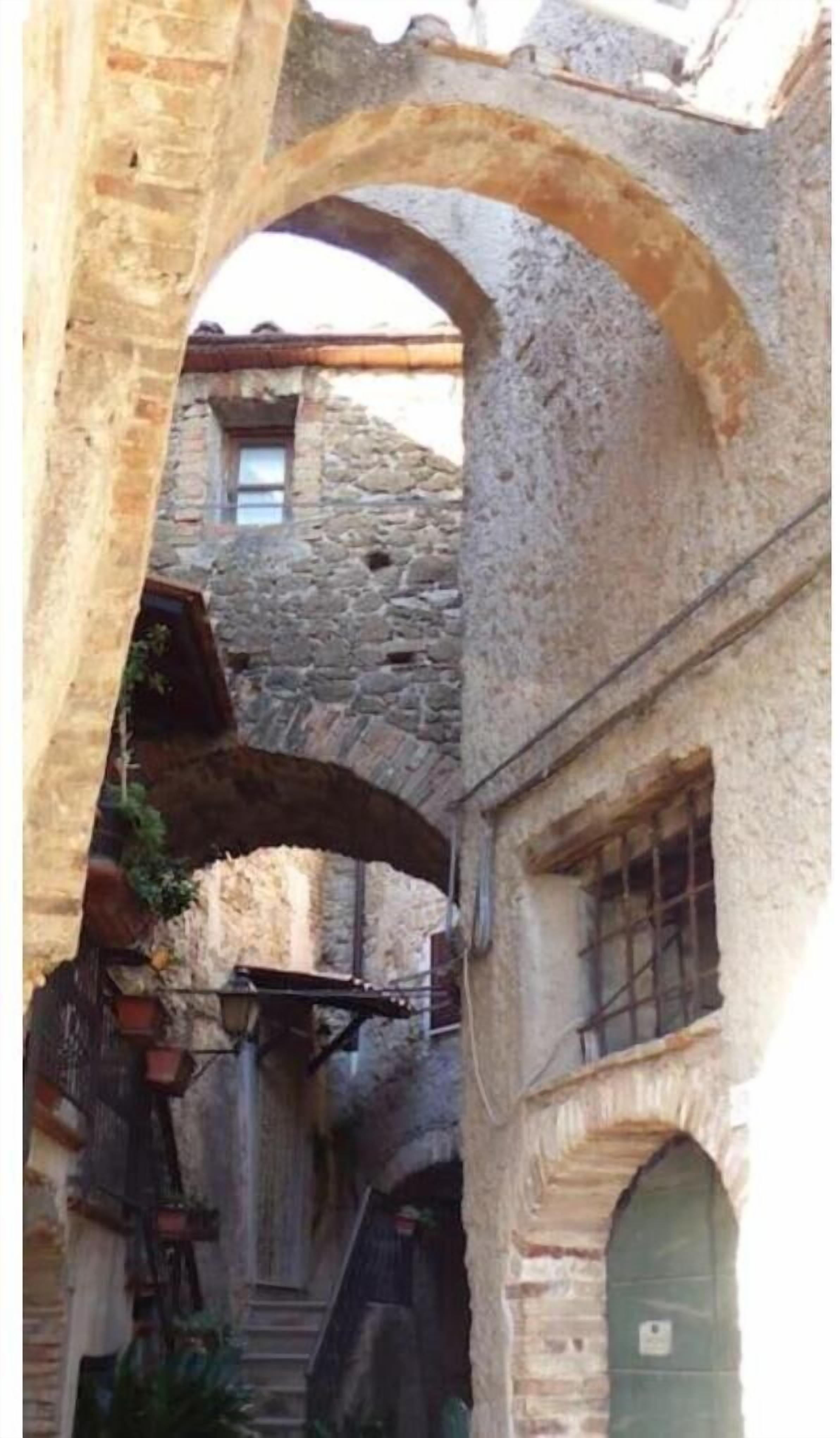 An arched stone corridor with a rustic stone building in the background.