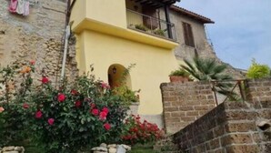 A two-story stone house with a yellow facade, a balcony, and a garden with red flowers. - Casa Romantica in un Borgo Antico in Sabina (Cicignano)