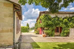 A courtyard with a gravel path, a building with red shutters, and a large planter. - Cottage 'Échappée Bel' with Shared Terrace, Shared Garden and Wi-Fi (arveyres)