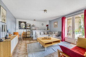A modern living room with a wooden coffee table, a sofa with red cushions, a dining area with chairs, a kitchen area with a microwave, and a large window with curtains. - Cottage 'Échappée Bel' with Shared Terrace, Shared Garden and Wi-Fi (arveyres)