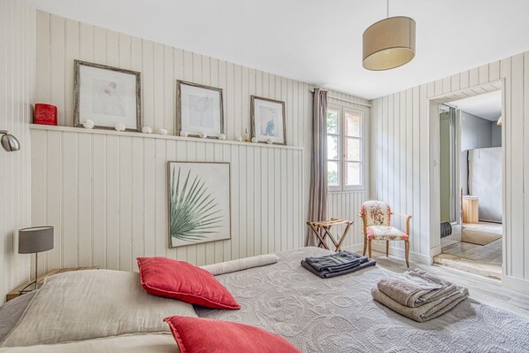 Un dormitorio con cama, almohadas, una silla, una estantería con cuadros enmarcados y una ventana.