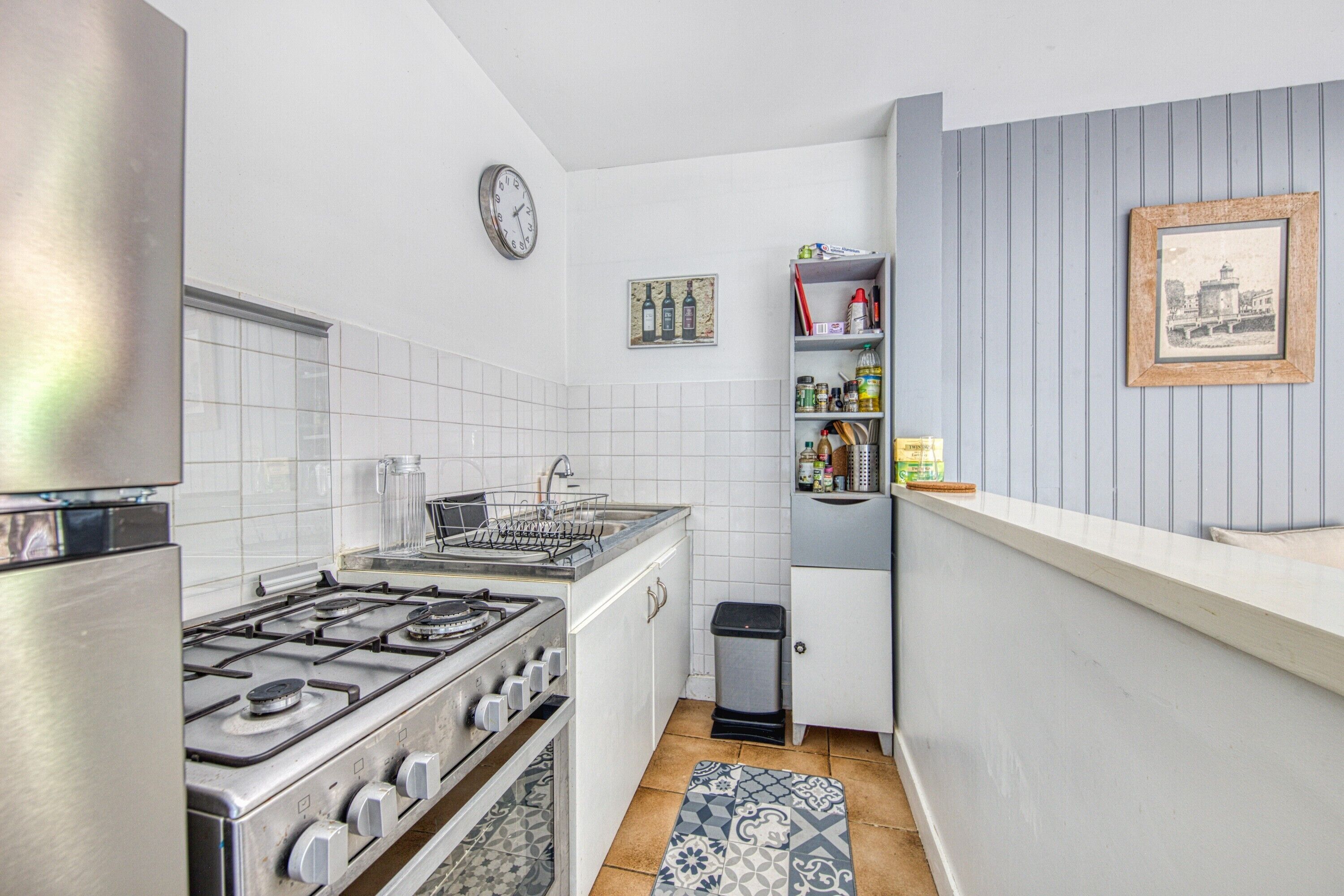 Une cuisine de taille réduite avec des meubles blancs, une cuisinière à gaz, un évier et une horloge au mur.