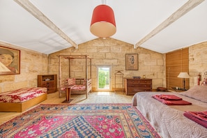 A bedroom with a stone wall, a large bed, a wooden chair, a small table, and a hanging light fixture.