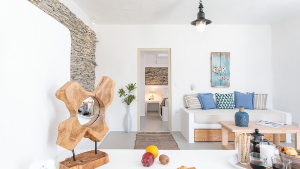 Ein modernes Wohnzimmer mit einem weißen Sofa, einer abstrakten Holzskulptur und einem Essbereich mit einer Platte voller Speisen und Getränke.
