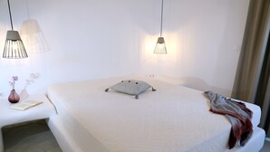 Een slaapkamer met een groot bed, bedlampjes, een nachtkastje met een vaas en boek, en een raam met gordijnen.