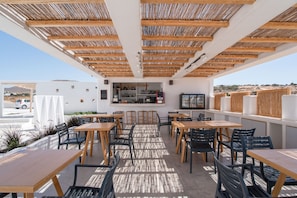 An outdoor seating area with wooden tables and chairs, a bar with shelves of bottles, and a view of the landscape. - Apartment at 'Irida Sunny Family' Hotel with Sea View, Wi-Fi and Air Conditioning (Naxos)
