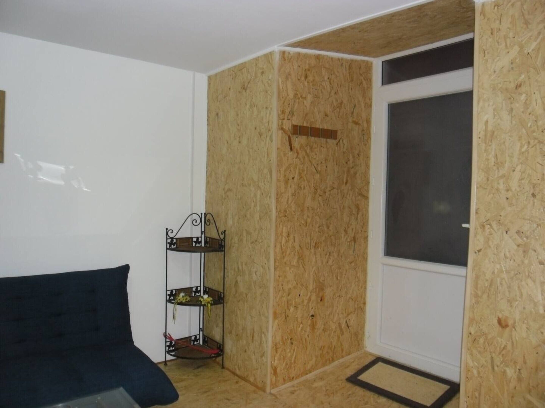 A room with a wooden wall panel, a metal shelf with flowers, a dark sofa, and a window.