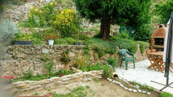 Un mur de soutènement en pierre, un jardin, une table et des chaises en bois, ainsi qu’un barbecue.