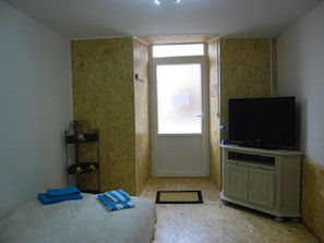 Une petite pièce comprenant un lit, un téléviseur posé sur un meuble, une porte donnant sur l’extérieur et une petite étagère avec quelques objets.