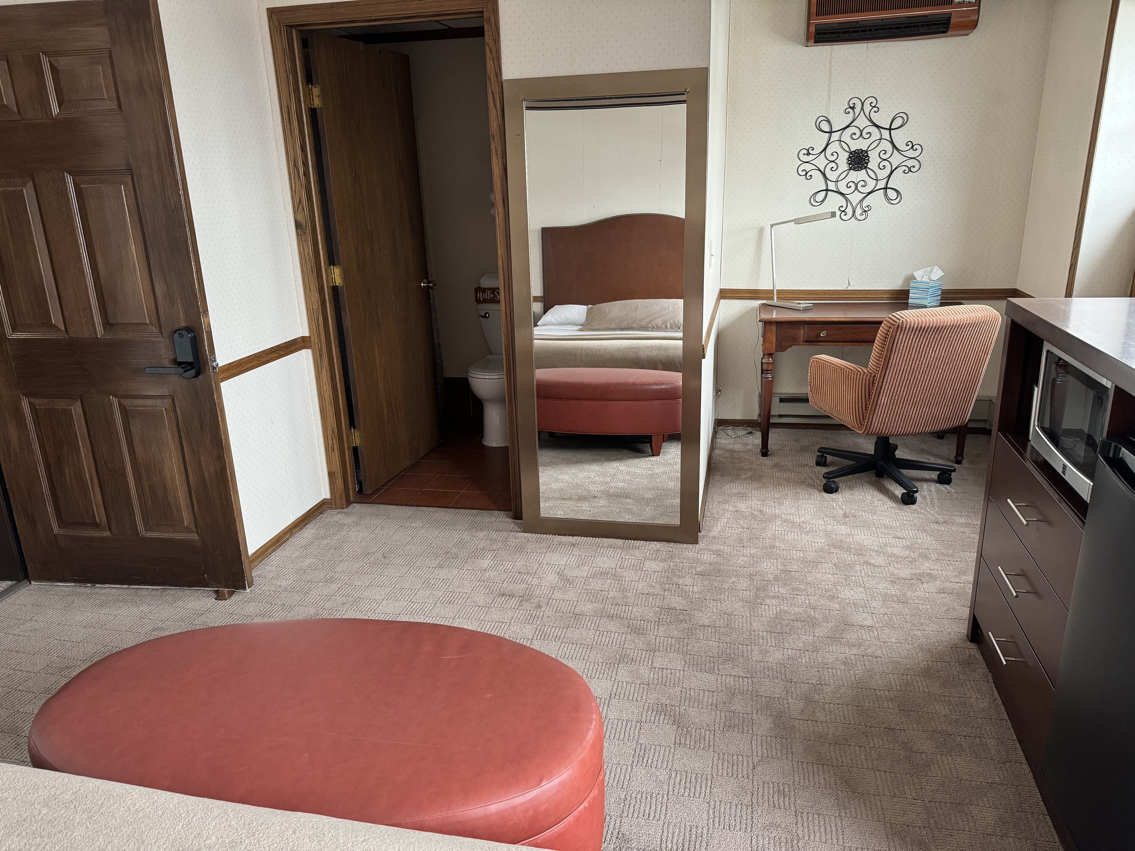 Habitación de hotel con puerta de madera, espejo, cama, escritorio, silla de oficina, televisor y mesita con caja de pañuelos.
