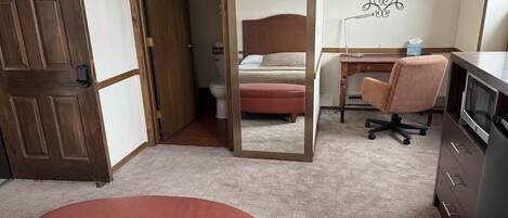 Habitación de hotel con puerta de madera, espejo, cama, escritorio, silla de oficina, televisor y mesita con caja de pañuelos.