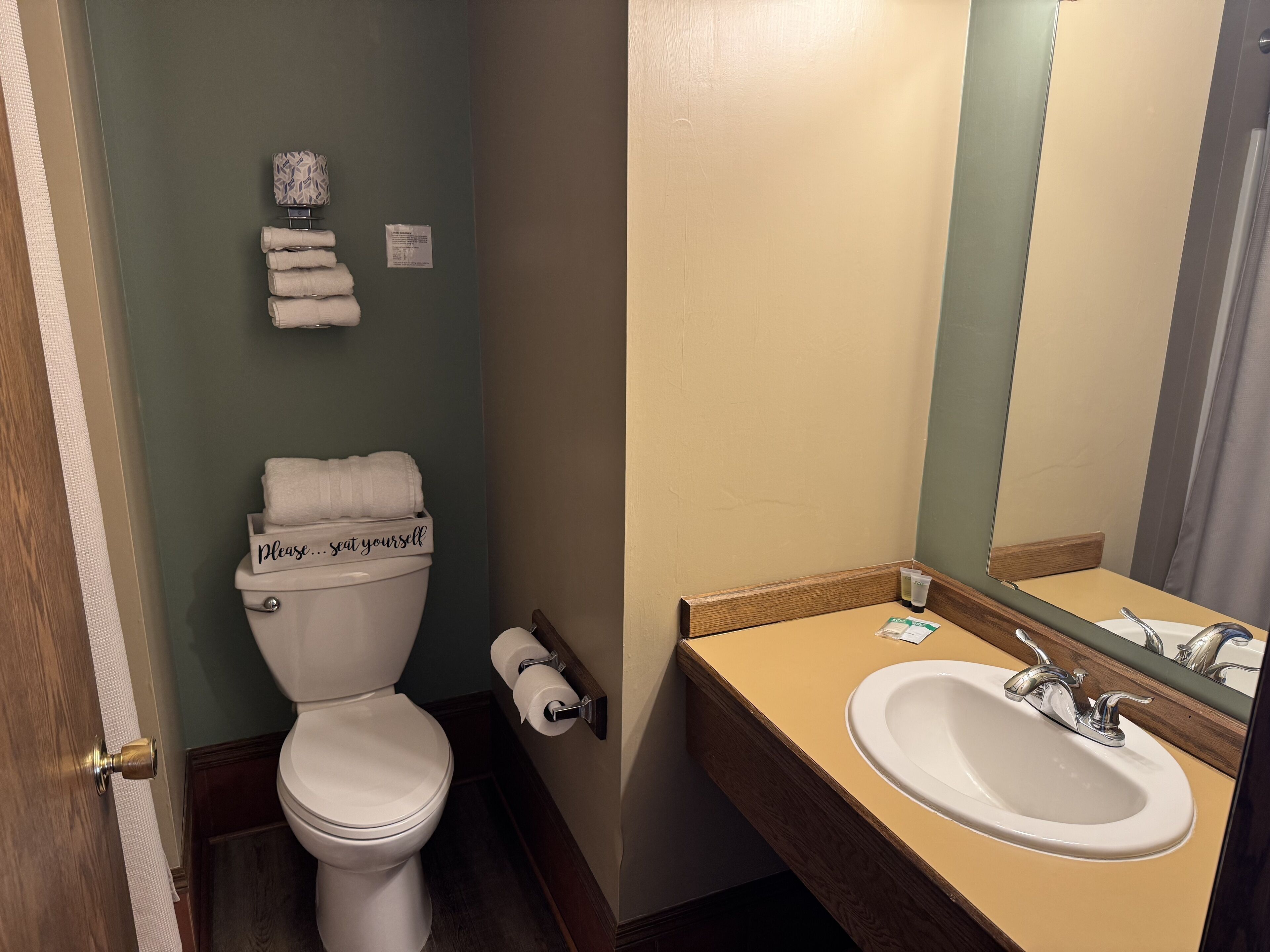 Un baño con inodoro, lavamanos y espejo. Hay toallas apiladas sobre el inodoro y un letrero que dice 'Por favor... tome asiento'.