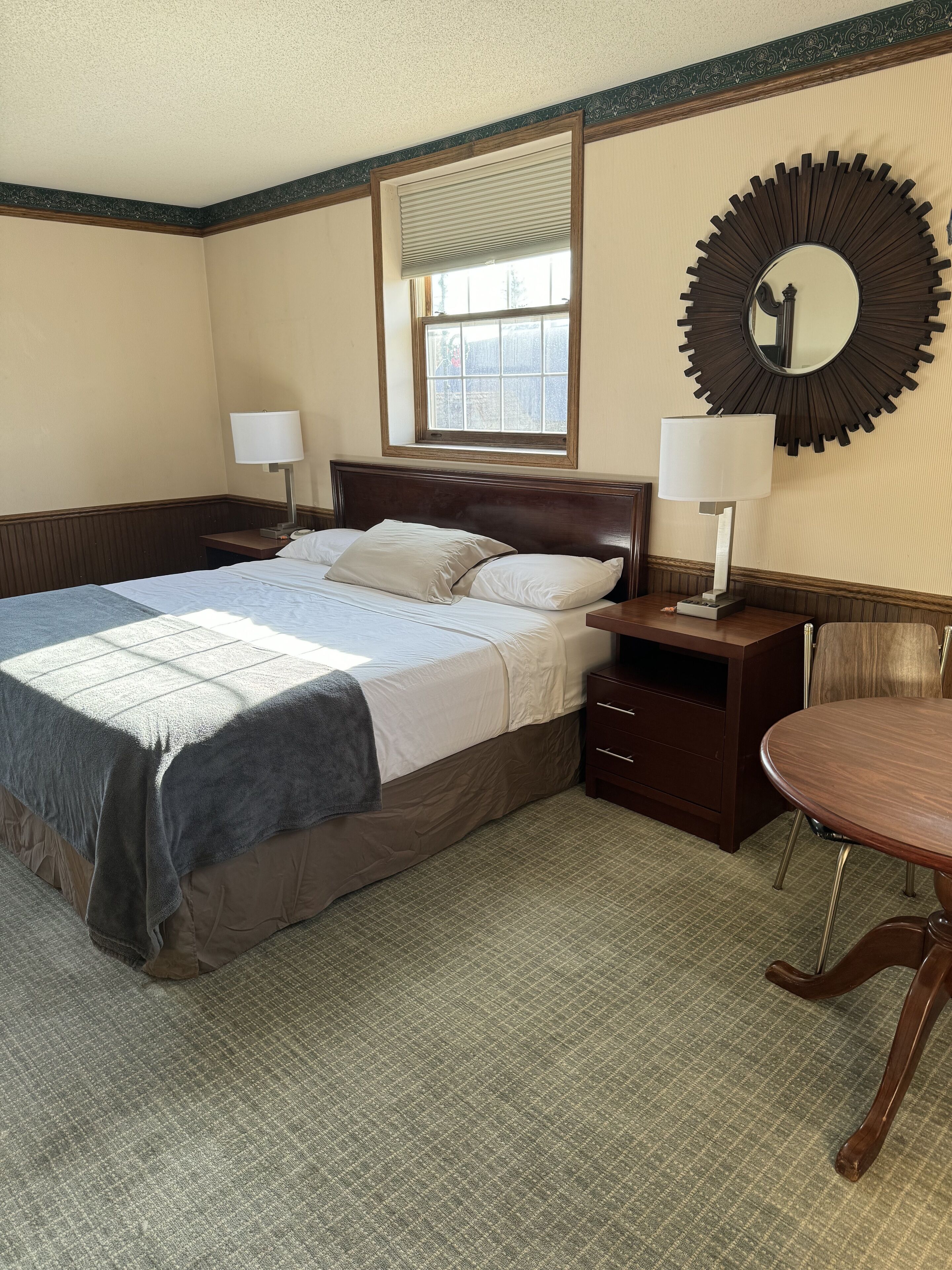 A hotel room with a large bed, two bedside tables with lamps, a round mirror, and a window with blinds.