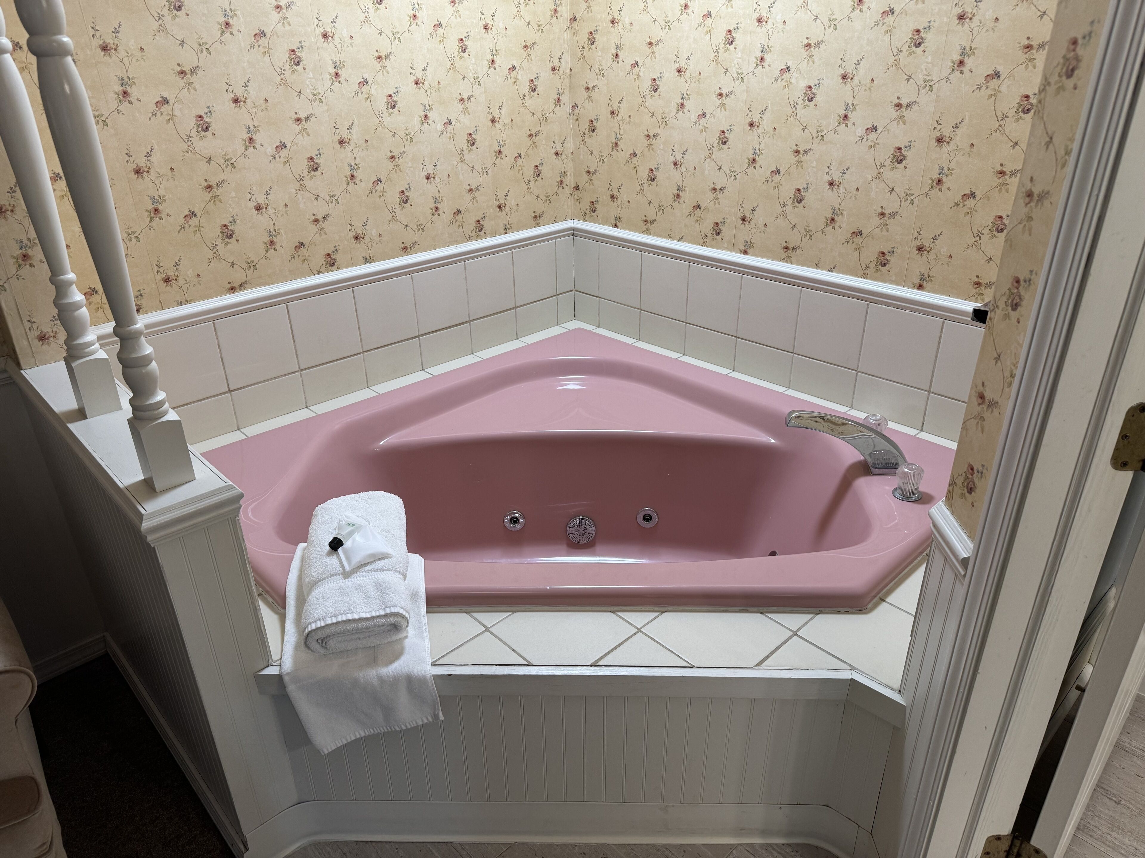 Eine rosafarbene Badewanne mit einem weißen Handtuch, weißen Fliesenwänden und Blumenpapier.