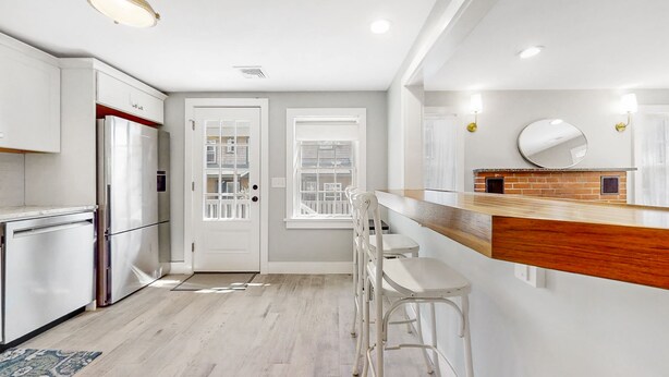 A modern kitchen with white cabinetry, a wooden countertop, and a brick backsplash. There are two white bar stools and a window with a view of the outside.