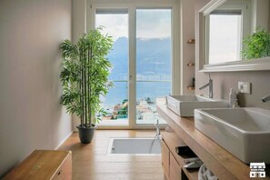 A modern bathroom with a large window, a potted plant, and a view of the city. - BELAGO by Design Studio (Bellano)