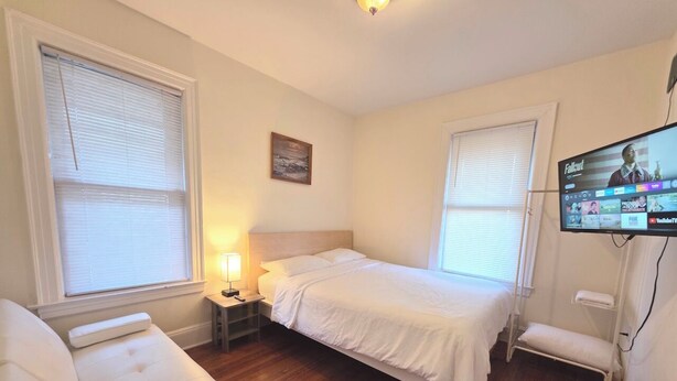 A bedroom with a bed, a bedside table with a lamp, a TV on the wall, and a window with blinds.
