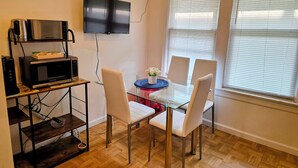 A dining area with a glass table, white chairs, a TV mounted on the wall, and a microwave on a shelf.