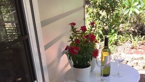 Una silla blanca al aire libre con un cojín colorido, una mesa blanca con una planta de flor roja en maceta, y dos copas de vino junto con una botella sobre la mesa.