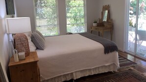 Un dormitorio con una cama grande, dos ventanas con persianas, una mesita de noche de madera con lámpara, un aparador con espejo y vistas al exterior con vegetación.
