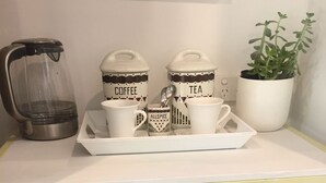 Una kitchenette con cafetera, dos tazas que dicen 'Café' y 'Té', una bandeja y una planta en maceta.