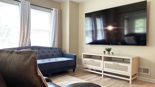 Un soggiorno con una grande TV a schermo piatto montata a parete, un divano blu scuro, un mobile TV bianco con cassetti e una finestra con tende.