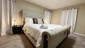 Un letto rifatto con testiera trapuntata, biancheria da letto bianca e un asciugamano piegato. È presente un comodino con una lampada e un piccolo cassetto. La stanza ha pareti e tappeto color beige, con tende bianche alle finestre.