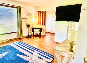 A room with a desk, chair, standing fan, and a large window with curtains.