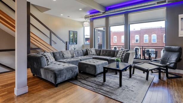 Ein modernes Wohnzimmer mit Ecksofa, Hocker, Couchtisch und Sessel. Eine Treppe mit Holzboden und Geländer ist zu sehen. Große Fenster bieten Blick auf ein Backsteingebäude.